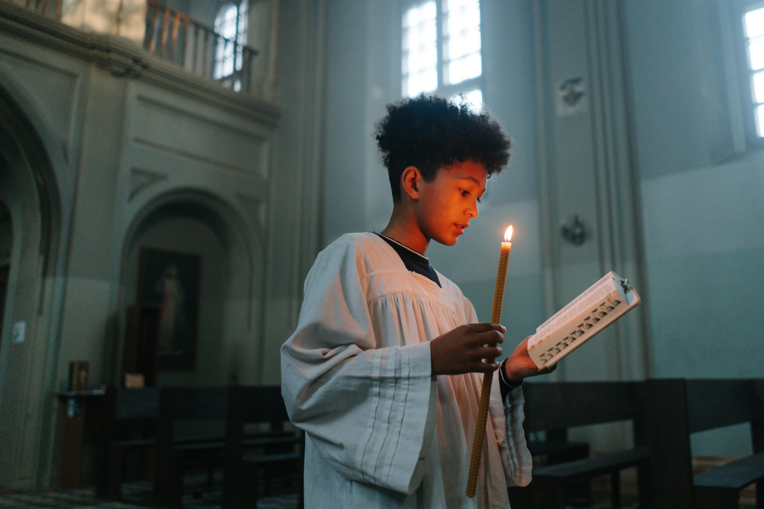 Child dressed in a robe holding a candle and book, reading inside a church with light streaming through windows.